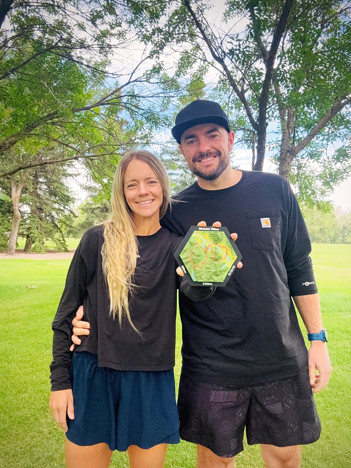 Cass and Weston de Winter standing together in a park, holding up a Sinister 50 km tile that reads 2399 m and 5:10:19.